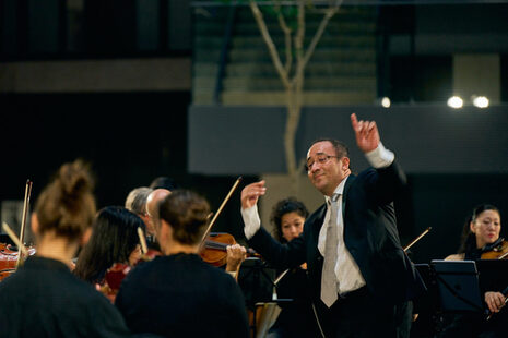 Un chef d'orchestre se tient devant l'orchestre, les bras levés.