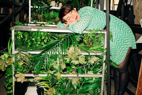 Una mujer con un vestido de punto verde menta se inclina sobre una estantería de plantas artificiales